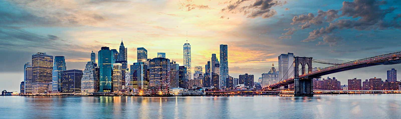 A panoramic cityscape with a bridge and skyline during sunset, viewed from across a river with the city s lights reflecting on its surface.