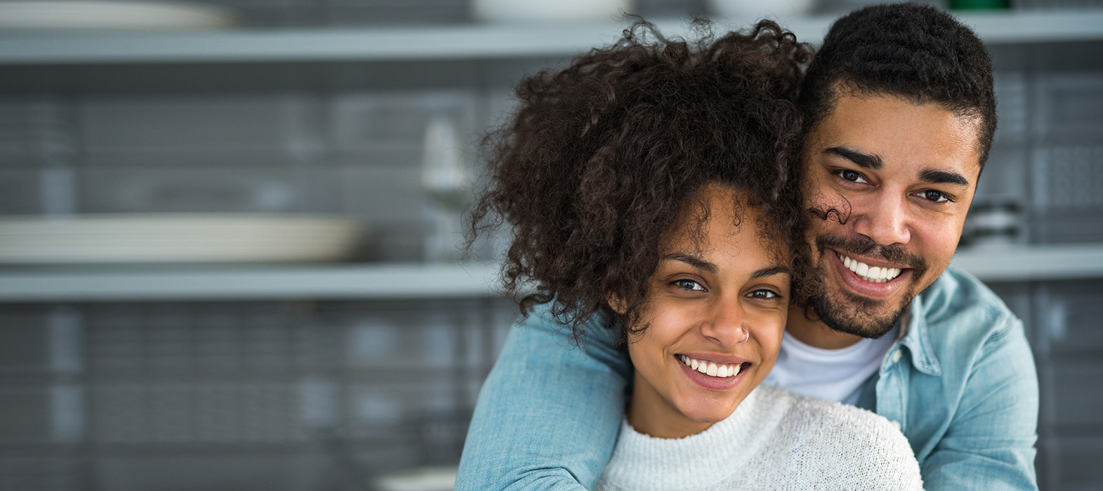 The image shows a man and a woman smiling at the camera, with the man s face partially obscured by the woman s arm. They appear to be in a modern indoor setting.