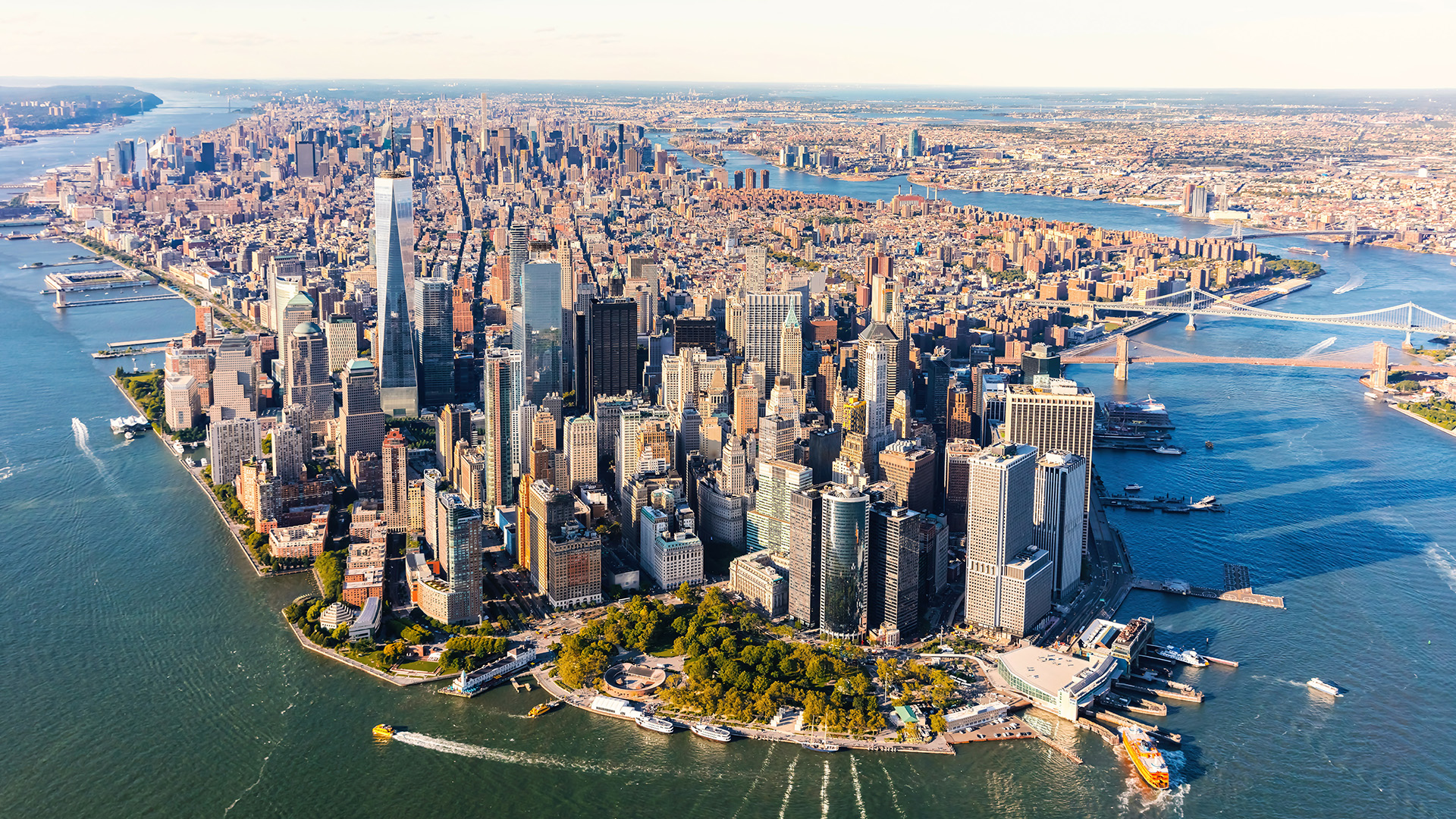 Aerial view of a city skyline with the Manhattan skyline, including iconic buildings such as the Empire State Building and the Chrysler Building, with the Statue of Liberty in the distance.