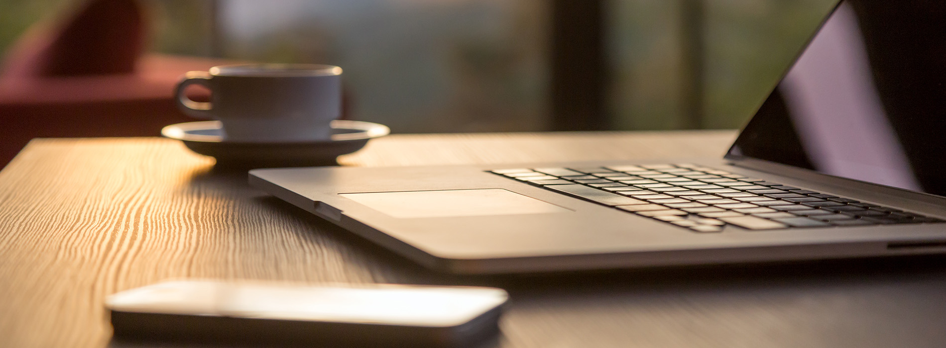 The image displays a laptop with an open screen, placed on a desk in front of a person who appears to be seated and working. There is a blurred view of the person's hands holding a cup of coffee, suggesting a casual work environment.