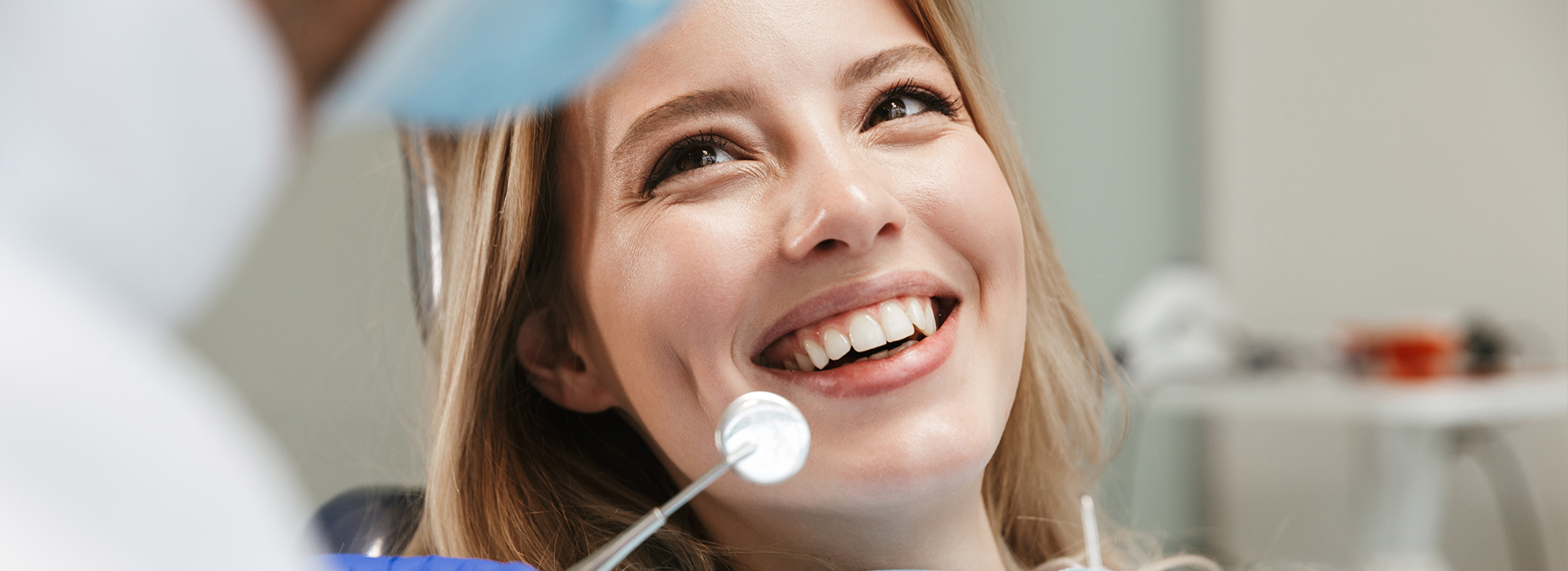 The image shows a woman sitting in a dental chair, receiving care from a dentist who is smiling and holding a mirror.