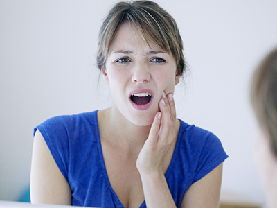 A woman with her mouth open, showing teeth, while looking at a mirror and touching her face.
